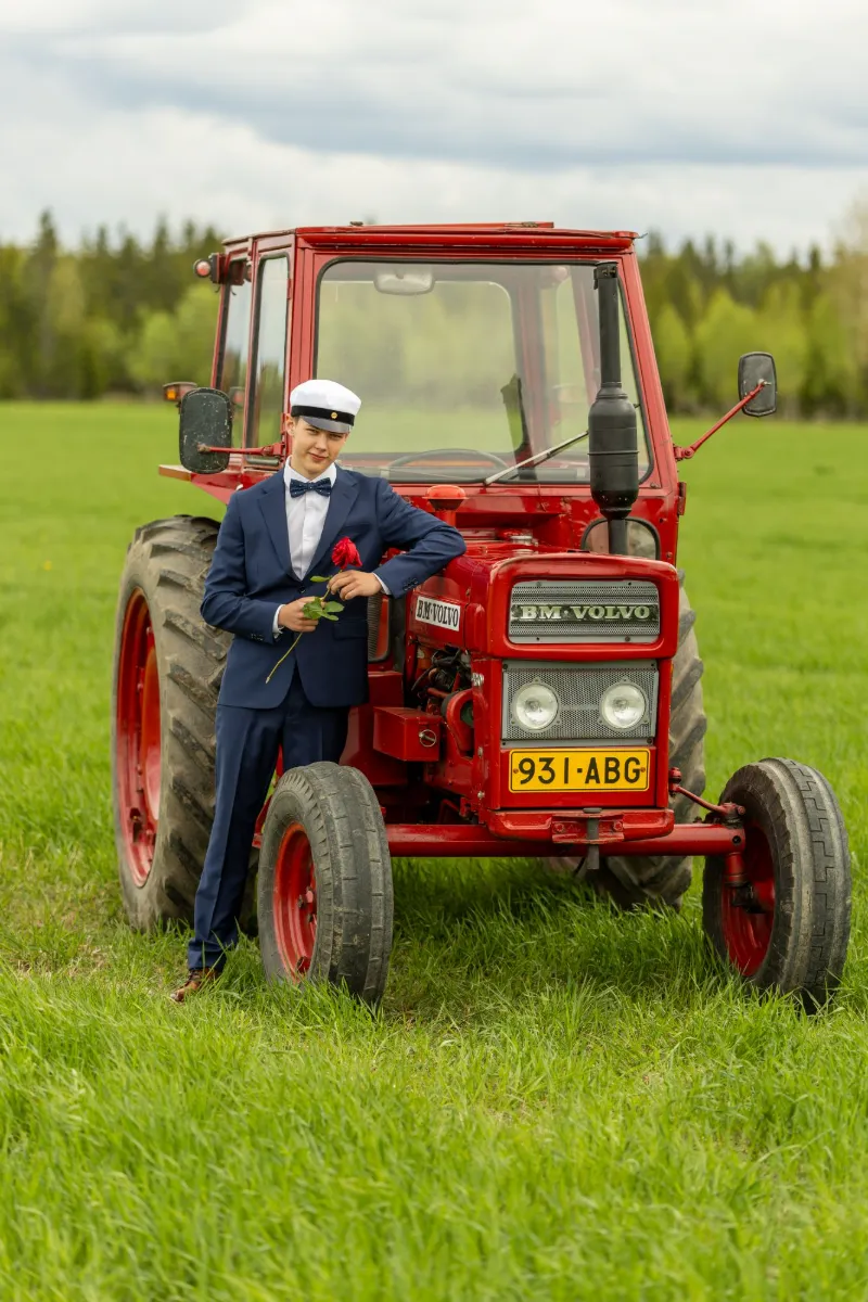 Ylioppilas tummansinisessä puvussa nojaa punaiseen BM Volvo -traktoriin vihreällä pellolla ruusu kädessä.