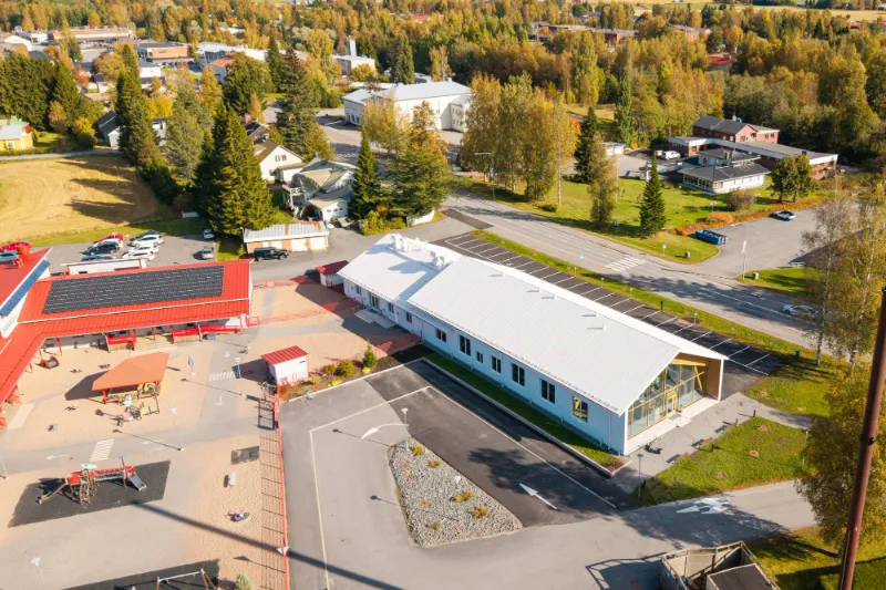Syksyinen ilmakuva valkoisesta uudisrakennuksesta, pysäköintialueesta ja viereisestä leikkipihasta.