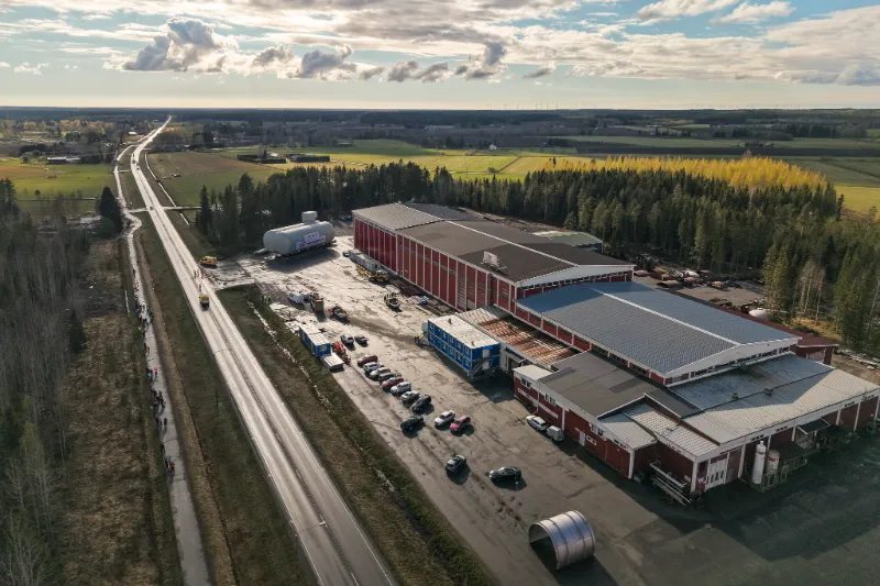Ilmakuva tehdasalueesta ja sen pihalla odottavasta suuresta säiliöstä. Maantien varrella yleisöä, taustalla pellot ja metsä.