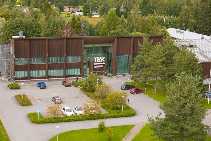 Ilmakuva puuverhoillusta toimitilarakennuksesta, jonka edessä on pysäköintialue, istutuksia ja mäntyjä.