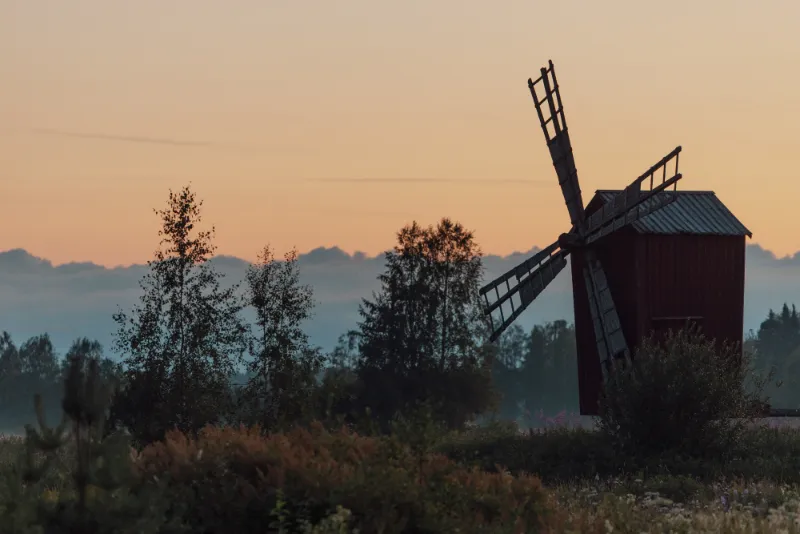 Punainen puinen tuulimylly ja koivuja siluettina oranssia aamutaivasta vasten.