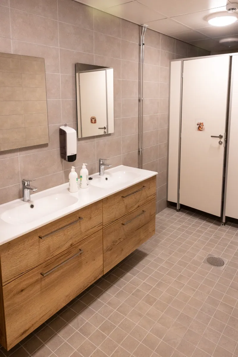Laatoitettu wc-tila, jossa on kaksoisallas puisella kaapistolla ja wc-koppien ovet.